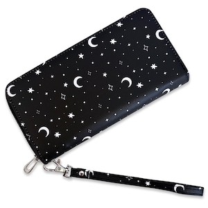 May include: A rectangular black wallet featuring a white celestial print of crescent moons, various star shapes, and small dots. The wallet includes a silver-toned metal zipper closure and a matching detachable wrist strap with the same pattern.