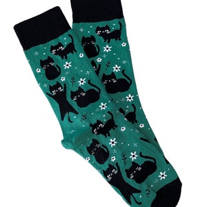 May include: A pair of green socks with a black cuff and a pattern of black cats and white flowers. The cats are in various poses, and the socks are made of a soft material.