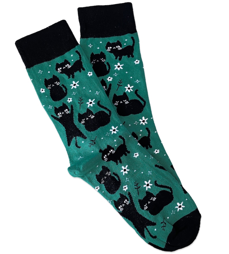 May include: A pair of green socks with a black cuff and a pattern of black cats and white flowers. The cats are in various poses, and the socks are made of a soft material.
