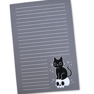 May include: A gray notepad with white horizontal lines. A black cat illustration sits atop a white skull, with small star accents. The notepad is rectangular and has a slight angle.