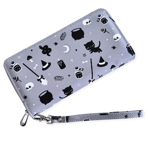 May include: A gray wallet with a wrist strap, featuring a repeating pattern of black witch-themed icons. The icons include cats, cauldrons, witch hats, potion bottles, skulls, and stars. The wallet has a silver zipper and a wrist strap.