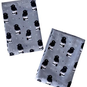 May include: Two gray microfiber cloths with a repeating pattern of black crows perched on white skulls. The design includes small stars. The cloths are folded and displayed on a white background.