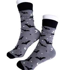 May include: A pair of gray socks with a black bat, moon, and star pattern. The socks have black cuffs, heels, and toes. The socks are displayed on white forms.