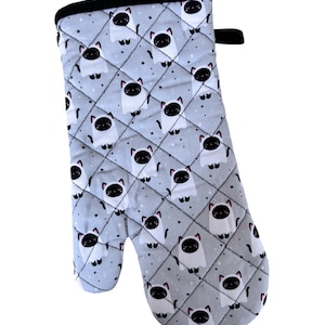 Peut inclure: Un gant de cuisine matelassé avec un motif répété de visages de chats blancs avec des accents noirs sur un fond gris clair. Le gant a une bordure noire et une boucle pour l'accrocher. Le motif comprend de petites étoiles.