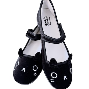 May include: A pair of black velvet Mary Jane style shoes with cat face embroidery. The shoes have a strap closure and small cat ear details. The inner sole is white with the text "EMS & Sprout".