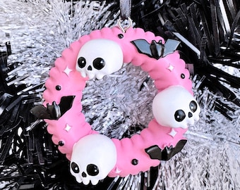 May include: A pink wreath-shaped ornament with three white skulls and three black bats. The wreath has a textured surface and is decorated with small star-shaped accents. A black ribbon is attached for hanging. The ornament is set against a backdrop of black and silver tinsel.