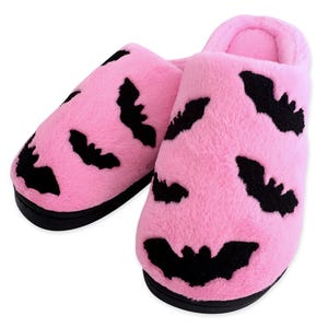 May include: A pair of pink plush slippers with black bat silhouettes. The slippers have a soft, fuzzy texture and a black sole. The bat design is repeated across the top of each slipper.
