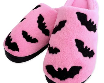 May include: A pair of pink plush slippers with black bat silhouettes. The slippers have a soft, fuzzy texture and a black sole. The bat design is repeated across the top of each slipper.