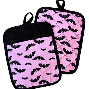 Peut inclure: Deux maniques matelassées avec un motif de chauve-souris noire sur fond rose. Les maniques ont une bordure noire et des boucles de suspension. Le motif de chauve-souris est répété sur le tissu rose, avec de petits accents d'étoiles blanches.
