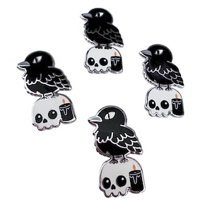 May include: Four black and white enamel pins featuring a black crow perched on a white skull with a black candle. The crow has a black body with white eyes and a black beak. The skull has a white body with black eyes and a black mouth. The candle is black with a yellow flame.