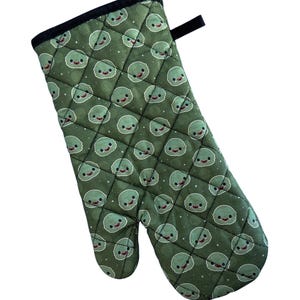May include: Green oven mitt with a quilted pattern and a repeating design of cartoon green faces with red mouths and white eyes.