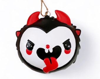 May include: A red and black Christmas ornament shaped like a devil with a white face and a red tongue sticking out. The ornament has a small loop at the top for hanging.