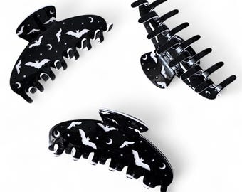 May include: Three black hair claw clips with a bat and moon design. The clips are made of a glossy material and feature white bat and crescent moon patterns. The clips are a stylish accessory for any hair type.