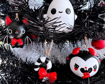 May include: A black Christmas tree decorated with Halloween-themed ornaments. The ornaments include a white ghost, a black bat with a red bow tie, a candy cane with a red bow, and a devil-faced ornament with red horns.