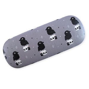 May include: A hard-shell, oval-shaped glasses case in a matte gray finish. The surface features a repeating pattern of black crows perched on white skulls, accompanied by small black candles and tiny white four-pointed star icons.