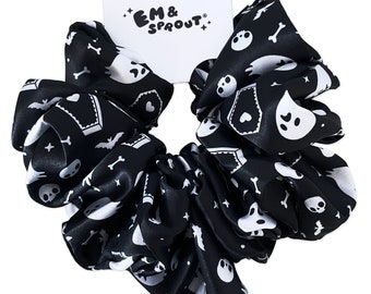 Puede incluir: Scrunchie de satén negro con estampado blanco de calaveras, huesos, murciélagos y fantasmas. El scrunchie tiene una etiqueta blanca con el texto "EM & SPROUT".