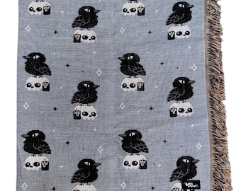 May include: A gray woven throw blanket featuring a repeating pattern of black crows perched atop white skulls with small black bottles. The blanket has a fringed edge and a small tag with text.
