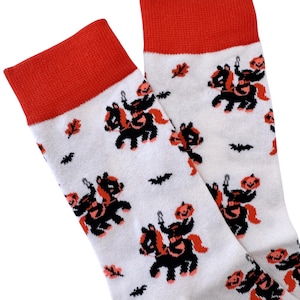 May include: A pair of white socks with a red cuff. The socks feature a repeating pattern of black and orange jack-o'-lantern figures riding black horses.