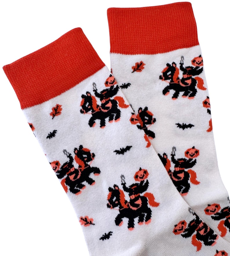 May include: A pair of white socks with a red cuff. The socks feature a repeating pattern of black and orange jack-o'-lantern figures riding black horses.