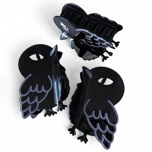 May include: Three black hair clips in the shape of owls with gray outlines and details. The clips are made of a glossy material and are arranged on a white background. The clips have a claw-like design to hold hair in place.
