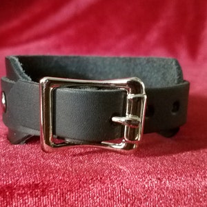 May include: A black leather cuff with a silver buckle. The cuff is adjustable and has a simple design.
