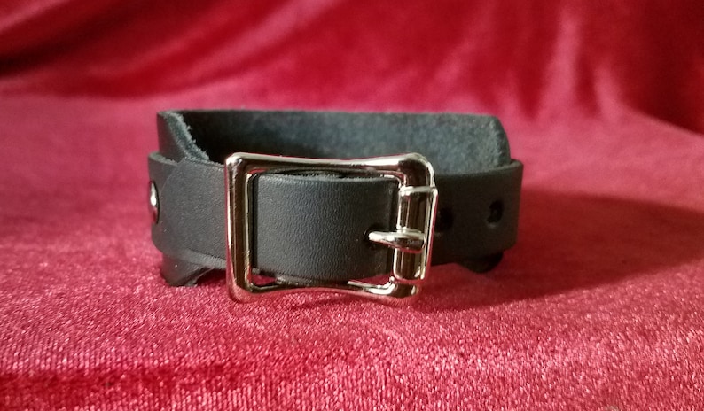 May include: A black leather cuff with a silver buckle. The cuff is adjustable and has a simple design.