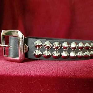 May include: Black leather bracelet with two rows of silver-coloured metal studs. The bracelet has a silver-coloured metal buckle. The background is a deep red colour.