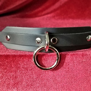 May include: Black leather choker with silver hardware, including a large ring and a buckle closure.