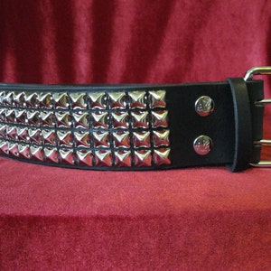May include: Black leather belt with three rows of silver pyramid studs. The belt has a silver buckle and two silver rivets near the buckle. The belt is displayed against a red background.