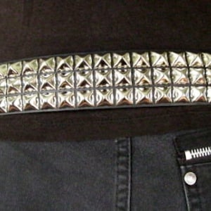 May include: A black leather belt with silver pyramid-shaped studs. The belt has three rows of studs and is worn around the waist.