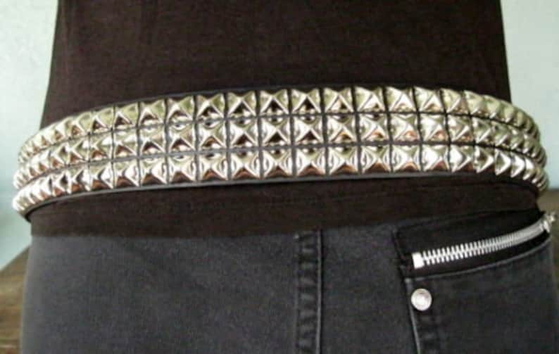 May include: A black leather belt with silver pyramid-shaped studs. The belt has three rows of studs and is worn around the waist.