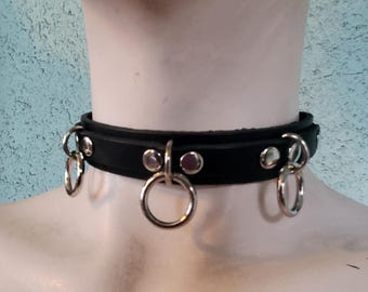 Può includere: Choker in pelle nera con anelli e borchie in metallo argentato.