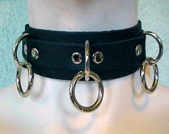 May include: A black leather choker with three silver rings. The choker is wide and has a smooth, matte finish. The rings are all different sizes and are attached to the choker with rivets.