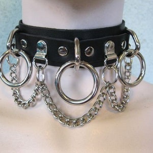 Puede incluir: Choker de cuero negro con múltiples anillos y cadenas plateadas. El choker tiene un diseño tachonado y es ajustable.
