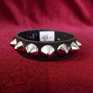 Puede incluir: Pulsera de cuero negro con tachuelas de pirámide plateadas. La pulsera tiene una etiqueta de talla que dice "ARE L".