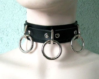May include: A black leather choker with three silver rings. The choker has silver studs along the edge.