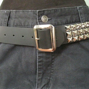 May include: A black leather belt with a silver buckle and pyramid-shaped studs. The belt is worn around the waist of a person wearing dark jeans.