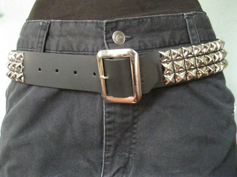 May include: A black leather belt with a silver buckle and pyramid-shaped studs. The belt is worn around the waist of a person wearing dark jeans.