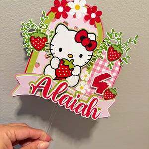 May include: A vibrant cake topper with Hello Kitty holding a strawberry. The design incorporates red strawberries, white flowers, and the name "Alaiah" in red script. A large red number 4 is also visible.