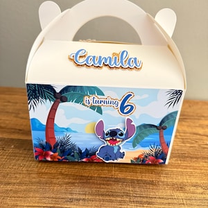 May include: A white cardboard party favor box with a blue Stitch character from Disney's Lilo & Stitch. The box has a handle and is decorated with palm trees, a beach scene, and the text "Camila is turning 6".