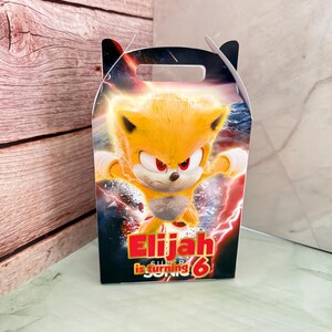 Puede incluir: Una caja de recuerdos de fiesta con un personaje de dibujos animados con pelaje amarillo y ojos rojos. La caja tiene el texto "Elijah is turning 6" en rojo y blanco. El fondo tiene un diseño de rayos azules y rojos.