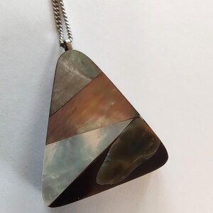 May include: A triangular pendant necklace featuring a mosaic of iridescent shell in shades of gray, copper, and white, set within a dark metal frame. The pendant hangs from a thin silver-toned metal chain with a fine link texture.
