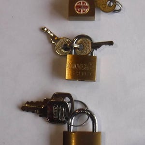 May include: Three brass padlocks with keys are displayed against a white background. The top lock has a red and white label. The middle lock is labeled "GUARD SECURITY". The bottom lock is labeled "ABUS" and "N 5/25".