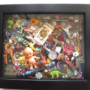 May include: A black framed shadow box filled with an assortment of colorful trinkets. The collection includes a giraffe, a baby doll, playing cards, a dinosaur, and a blue crayon. The items are arranged in a visually appealing, eclectic display.