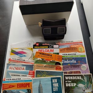 May include: A vintage View-Master set with a black viewer and a collection of colorful reels. The box is gray and black. Reels feature destinations like Europe, New York City, and Disney's Cinderella.