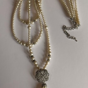 May include: Elegant multi-strand pearl necklace with a silver-toned floral pendant and tassel. The necklace features small, round pearls and decorative silver accents. A classic piece of jewelry.
