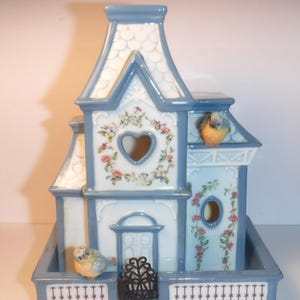 May include: A decorative ceramic birdhouse in shades of blue and white, adorned with floral accents and heart-shaped cutouts. Two small bird figurines perch on the roof and balcony. The birdhouse has a small decorative fence.