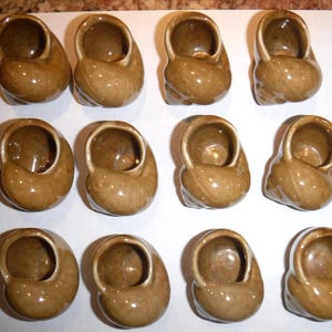 May include: A collection of twelve ceramic escargot dishes. Each dish is a light brown color, shaped like a snail shell, with a circular opening at the top. The dishes are arranged in three rows of four.