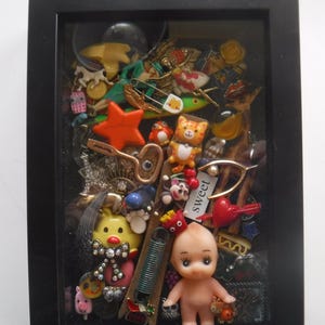 May include: A framed shadow box filled with an assortment of small, colorful trinkets. Includes a baby doll, a star, a cat figurine, and a tag that says "sweet". The frame is black.