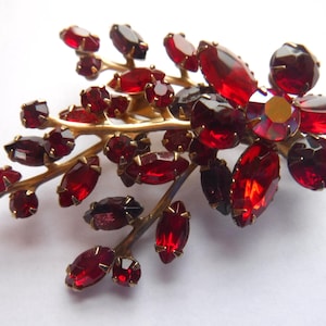 May include: A vintage brooch in the shape of a floral branch. The brooch features a gold-tone metal base with red, oval-shaped rhinestones and a central flower with a gold center. The rhinestones vary in size and shade.